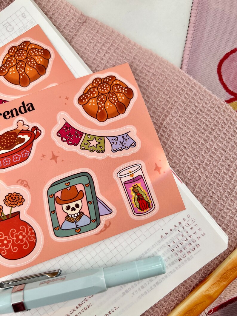 Op de afbeelding: Een set Day of the Dead-thema stickers met een suiker schedel, een pan de muerto, een kaars, een bloempot en een banner. De stickers zijn op een roze achtergrond.