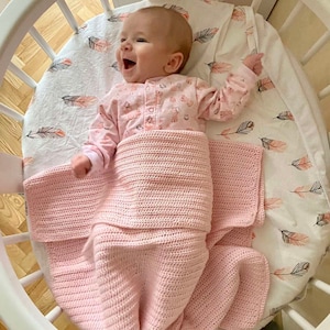 May include: A baby in a pink and white patterned onesie is lying in a white round crib. The baby is smiling and has a pink crocheted blanket wrapped around them. The crib has a white wooden frame and a white mattress.