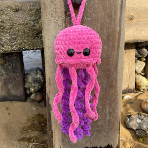 May include: A handmade, plush jellyfish toy in shades of pink and purple. The jellyfish has a round head with black button eyes and a loop for hanging. The tentacles are a mix of pink and purple, creating a whimsical ocean-themed decoration.