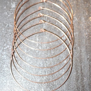 May include: A collection of copper-colored metal choker necklaces. Each necklace is a simple, thin, circular band. The necklaces are arranged in a layered pattern, with each one slightly larger than the last. The metal has a warm, reddish-brown hue.
