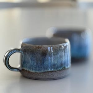 Könnte beinhalten: Zwei Keramikbecher mit kurzer, breiter zylindrischer Form und einem Henkel. Die Außenseite ist mit einer reaktiven Glasur in Blau- und Grautönen versehen, die am Boden in eine texturierte, erdfarbene braune Basis übergeht.