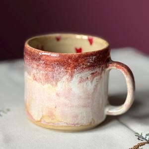 Może przedstawiać: Ręcznie robiony ceramiczny kubek z zaokrąglonym uchwytem. Kubek ma gradient kolorów, przechodzący od głębokiej czerwonej krawędzi do miękkiej różowo-białej podstawy. Małe czerwone serca zdobią wnętrze.