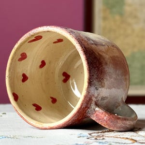 Może przedstawiać: Ręcznie robiony ceramiczny kubek w czerwono-kremowej kolorystyce. Wnętrze zdobią małe czerwone serca. Kubek ma zaokrąglony kształt z uchwytem i spoczywa na wzorzystej powierzchni.
