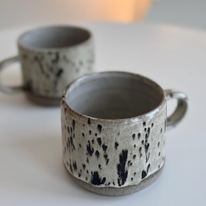Może przedstawiać: Dwa ceramiczne kubki z nakrapianymi czarno-szarymi wzorami. Kubki mają cylindryczny kształt z uchwytem i jasnoszarym wnętrzem. Kubki są ręcznie robione i nadają się do kawy lub herbaty.