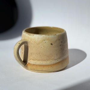Op de afbeelding: Een handgemaakte keramische mok met een afgeronde body en een klein handvat. De mok heeft een beige kleur met donkere spikkels en een lichtere band aan de basis. De binnenkant is lichtbruin. De mok staat op een wit oppervlak.