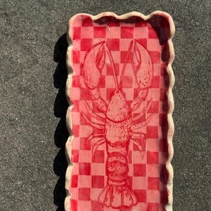 Könnte beinhalten: Ein rechteckiges Keramiktablett mit rot-weißem Schachbrettmuster und einer Hummerillustration. Das Tablett hat einen gewellten Rand und eignet sich zum Servieren von Vorspeisen oder zum Präsentieren kleiner Gegenstände.
