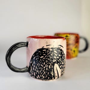 May include: Two ceramic mugs. The front mug is light pink with a black handle and a black and white design. The inside of the mug is red. The second mug is out of focus, with a yellow and orange design.