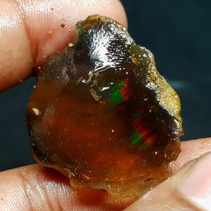 May include: A raw opal gemstone with a dark brown base colour. The opal displays flashes of green, red, and purple, and is held in a person's hand. The stone has a rough, uneven texture and a golden-brown edge.