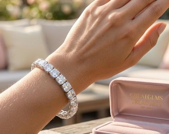 May include: A dazzling bracelet featuring multiple square-cut, clear gemstones set in a silver or white metal band. The bracelet is displayed on a wrist, with a pink velvet jewelry box in the background. The box has gold lettering that reads "GREAT GEMS TREASURES (EST. 2014)".