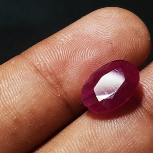 May include: A faceted, oval-shaped ruby gemstone. The ruby is a deep, rich red color with a brilliant, reflective surface. The gemstone is held in a hand, with the background out of focus.