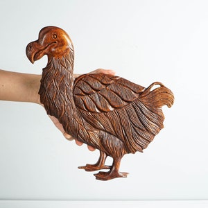 Puede incluir: Escultura de madera tallada de un pájaro dodo, con detalles de plumas y cuerpo. La escultura es de color marrón cálido, con un acabado liso. El ave se representa de pie.