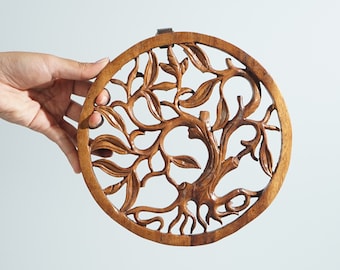 May include: A circular wooden wall hanging featuring an intricate, hand-carved design of a tree with sprawling branches and leaves. The natural brown wood grain is visible throughout the piece, which is framed by a solid circular border.
