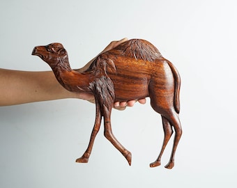 May include: A dark brown, hand-carved wooden camel figurine featuring detailed textures on the neck, hump, and legs. The sculpture depicts the animal in a walking stance with a smooth, polished finish that highlights the natural wood grain patterns.