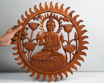 May include: A circular brown wooden wall carving featuring a central seated Buddha figure in a meditative pose. The figure is surrounded by carved lotus flowers and leaves, all enclosed within a sunburst-style frame with pointed, flame-like edges.
