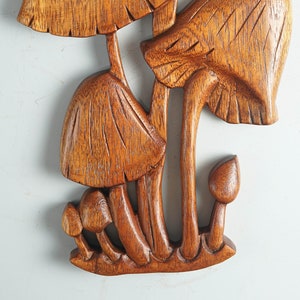 Op de afbeelding: Een bruin houten wandsculptuur met een groep paddenstoelen van verschillende groottes. Het stuk toont gedetailleerde houtnerven, met ingesneden lijnen op de hoeden en stelen van de paddenstoelen, die allemaal verbonden zijn aan een basis.