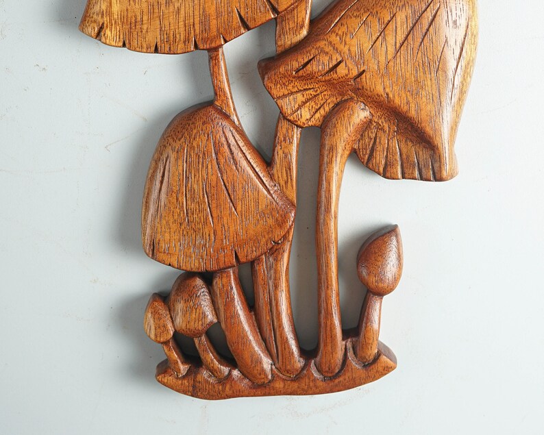 Op de afbeelding: Een bruin houten wandsculptuur met een groep paddenstoelen van verschillende groottes. Het stuk toont gedetailleerde houtnerven, met ingesneden lijnen op de hoeden en stelen van de paddenstoelen, die allemaal verbonden zijn aan een basis.