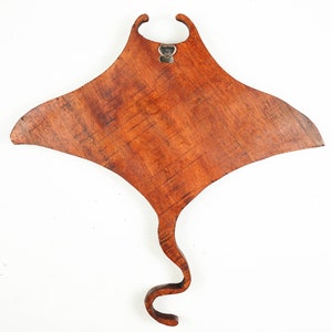 Op de afbeelding: Een houten wanddecoratie in de vorm van een mantarog. De rog is gemaakt van donkerbruin hout met een natuurlijke houtnerf. Hij heeft een metalen haak aan de bovenkant om op te hangen.