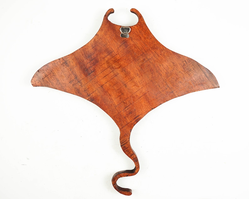 Op de afbeelding: Een houten wanddecoratie in de vorm van een mantarog. De rog is gemaakt van donkerbruin hout met een natuurlijke houtnerf. Hij heeft een metalen haak aan de bovenkant om op te hangen.