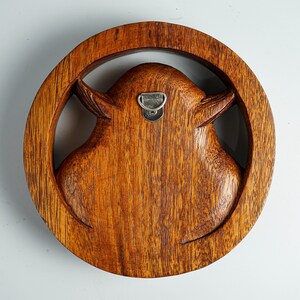 May include: A circular brown wooden wall carving featuring a stylized animal head silhouette centered within a thick wooden ring. The piece displays a visible wood grain texture and includes a silver metal D-ring hanger attached to the back.