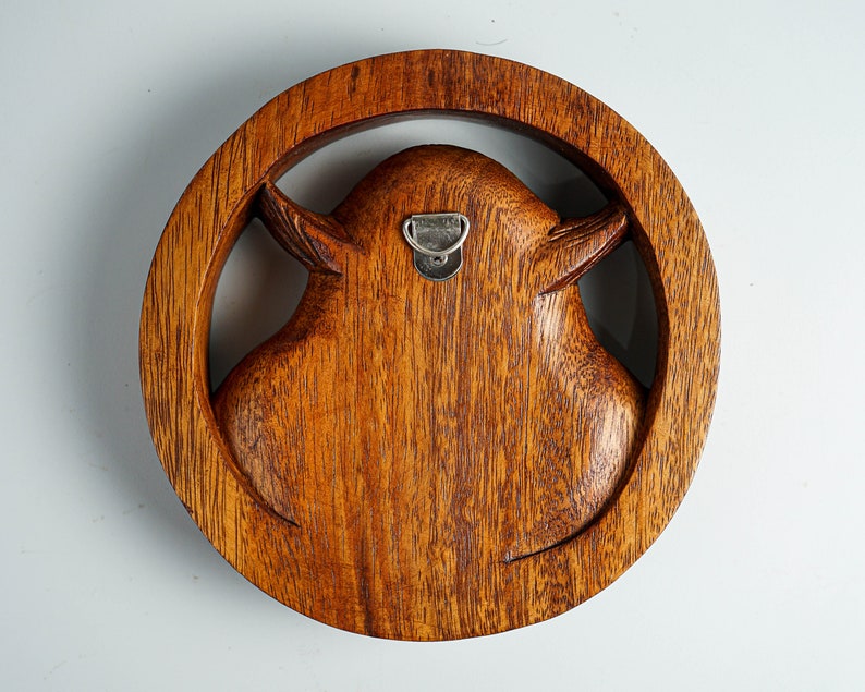 May include: A circular brown wooden wall carving featuring a stylized animal head silhouette centered within a thick wooden ring. The piece displays a visible wood grain texture and includes a silver metal D-ring hanger attached to the back.
