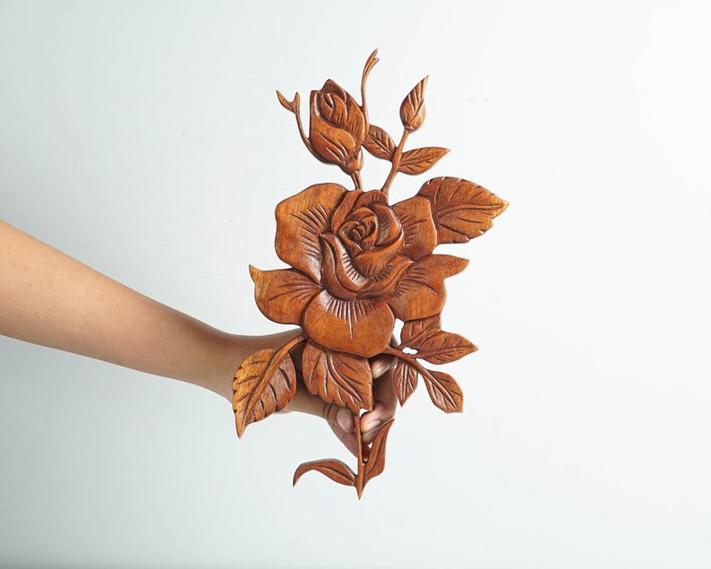 K&ouml;nnte beinhalten: Eine mittelbraune Holzwandschnitzerei, die eine detaillierte Rose mit einer zentralen Bl&uuml;te, einer kleineren Rosenknospe und mehreren gezackten Bl&auml;ttern zeigt. Die Holzmaserung ist auf dem gesamten St&uuml;ck sichtbar, das aufwendige Reliefschnitzereien aufweist.