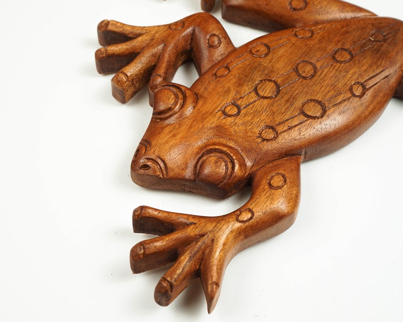 May include: A brown wooden frog carving featuring a smooth, polished finish and detailed incised circular patterns along its back and head. The sculpture displays distinct anatomical features including splayed toes and prominent eye ridges.