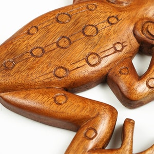 May include: A carved brown wooden frog figurine featuring a smooth, polished finish and a series of etched circular patterns along its back and legs. The wood grain is visible throughout the surface, highlighting the natural texture of the material.