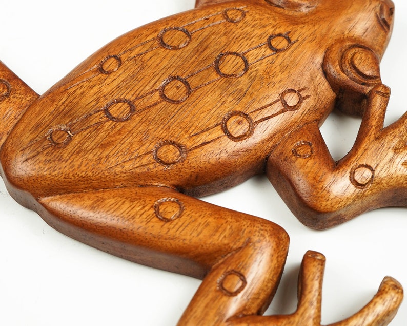 May include: A carved brown wooden frog figurine featuring a smooth, polished finish and a series of etched circular patterns along its back and legs. The wood grain is visible throughout the surface, highlighting the natural texture of the material.