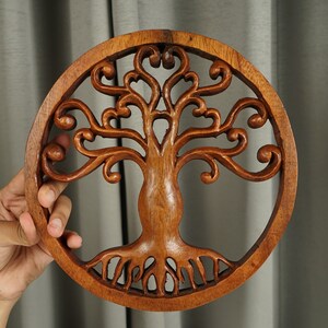 May include: A round wooden wall decor featuring a carved tree of life design. The tree has intricate branches and roots, and the wood is a rich brown color.
