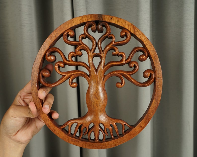 May include: A round wooden wall decor featuring a carved tree of life design. The tree has intricate branches and roots, and the wood is a rich brown color.