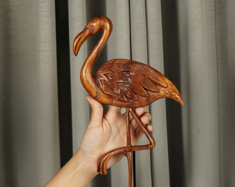Pu&ograve; includere: Una scultura in legno di un fenicottero rosa, in piedi su una sola zampa con la testa alta. La scultura &egrave; dettagliata con le piume e ha una finitura in legno naturale.