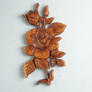 K&ouml;nnte beinhalten: Eine braune Holzwandschnitzerei, die eine detaillierte Rose mit einer Knospe, mehreren gezackten Bl&auml;ttern und einem Stiel darstellt. Die Holzmaserung ist im gesamten Relief sichtbar und zeigt aufwendig geschnitzte Texturen auf den Bl&uuml;tenbl&auml;ttern.