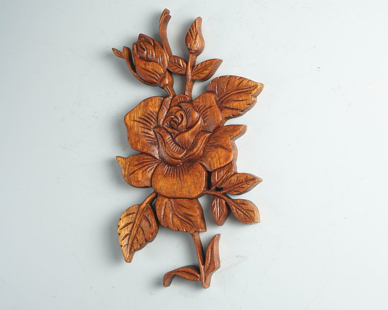 K&ouml;nnte beinhalten: Eine braune Holzwandschnitzerei, die eine detaillierte Rose mit einer Knospe, mehreren gezackten Bl&auml;ttern und einem Stiel darstellt. Die Holzmaserung ist im gesamten Relief sichtbar und zeigt aufwendig geschnitzte Texturen auf den Bl&uuml;tenbl&auml;ttern.