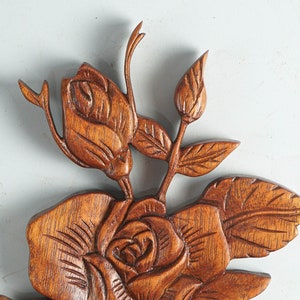 K&ouml;nnte beinhalten: Eine braune Holzschnitzerei f&uuml;r die Wand mit einem detaillierten Blumenmotiv, bestehend aus einer gro&szlig;en bl&uuml;henden Rose in der Mitte, begleitet von einer kleineren Rosenknospe und einer geschlossenen Knospe, verbunden durch St&auml;ngel und Bl&auml;tter.