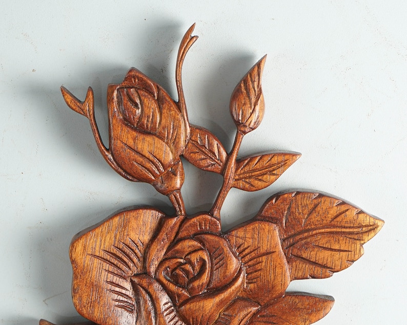 K&ouml;nnte beinhalten: Eine braune Holzschnitzerei f&uuml;r die Wand mit einem detaillierten Blumenmotiv, bestehend aus einer gro&szlig;en bl&uuml;henden Rose in der Mitte, begleitet von einer kleineren Rosenknospe und einer geschlossenen Knospe, verbunden durch St&auml;ngel und Bl&auml;tter.