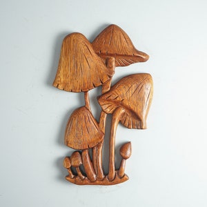 Op de afbeelding: Een houten wandsculptuur die een groep paddenstoelen van verschillende groottes weergeeft. Het stuk heeft een rijke bruine houtnerfafwerking met gedetailleerde verticale lijnen in de hoeden en stelen, wat zorgt voor een textuurrijk reli&euml;f.