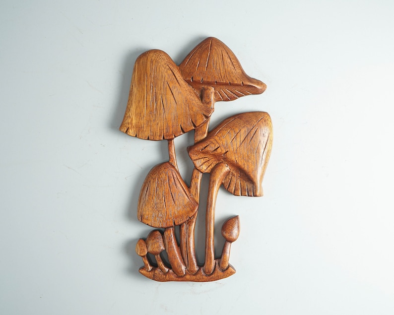 Op de afbeelding: Een houten wandsculptuur die een groep paddenstoelen van verschillende groottes weergeeft. Het stuk heeft een rijke bruine houtnerfafwerking met gedetailleerde verticale lijnen in de hoeden en stelen, wat zorgt voor een textuurrijk reli&euml;f.