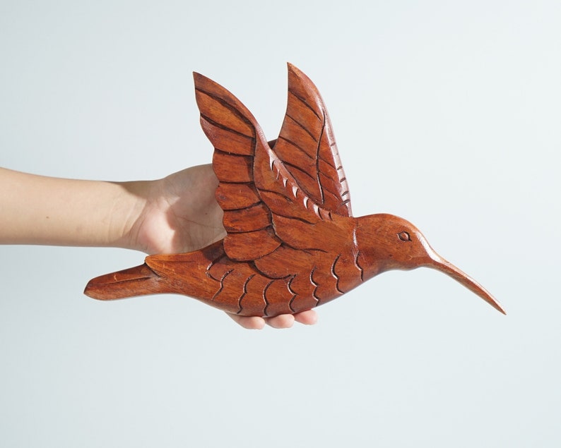 May include: A hand holds a wooden hummingbird sculpture with intricate details. The sculpture is painted in shades of brown and has a long, thin beak.