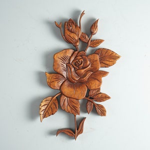 K&ouml;nnte beinhalten: Eine braune Holzwandschnitzerei, die eine detaillierte Rose mit einer zentralen bl&uuml;henden Bl&uuml;te, einer kleineren Rosenknospe an der Spitze und mehreren gezackten Bl&auml;ttern am zentralen Stiel zeigt, mit sichtbarer nat&uuml;rlicher Holzmaserung.