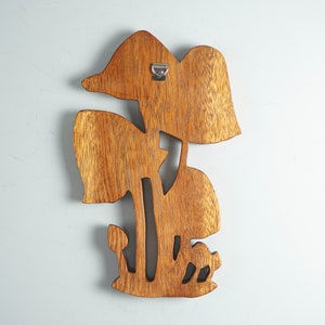 Op de afbeelding: Een bruine houten wanddecoratie met een uitgesneden ontwerp van meerdere paddenstoelen van verschillende groottes. Het stuk heeft een zichtbare houtnerfstructuur en bevat een zilverkleurige metalen D-ringhanger aan de achterkant voor montage.