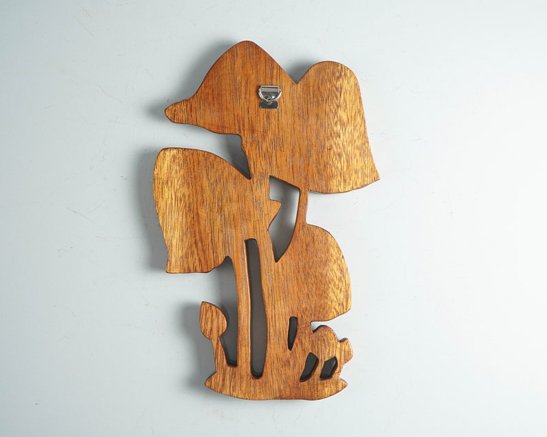 Op de afbeelding: Een bruine houten wanddecoratie met een uitgesneden ontwerp van meerdere paddenstoelen van verschillende groottes. Het stuk heeft een zichtbare houtnerfstructuur en bevat een zilverkleurige metalen D-ringhanger aan de achterkant voor montage.