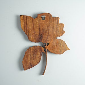 May include: A wooden wall decor in the shape of a rose with a brown stain finish. The rose has a single stem and leaves. The rose is attached to a white wall with a metal hook.