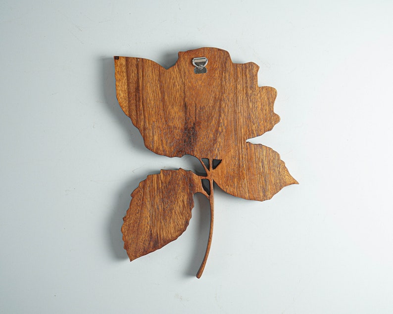 May include: A wooden wall decor in the shape of a rose with a brown stain finish. The rose has a single stem and leaves. The rose is attached to a white wall with a metal hook.