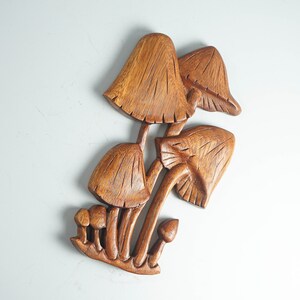 Op de afbeelding: Een bruin houten wandsculptuur met een groep paddenstoelen van verschillende groottes. Het stuk toont gedetailleerde houtnerven en ingesneden lijnen op de hoeden en stelen, wat een groep paddenstoelen weergeeft die uit een basis groeien.