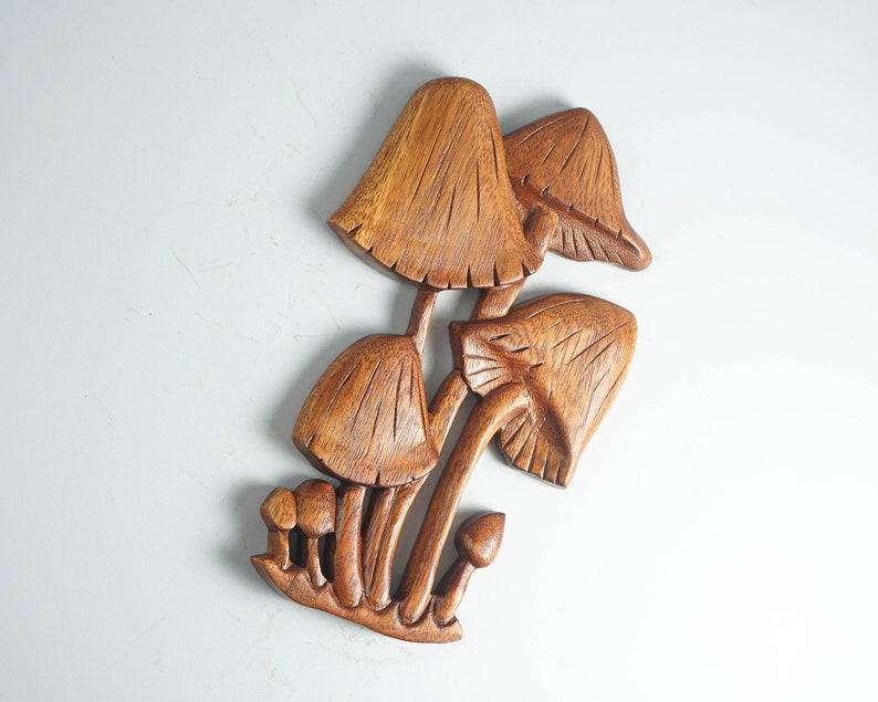 Op de afbeelding: Een bruin houten wandsculptuur met een groep paddenstoelen van verschillende groottes. Het stuk toont gedetailleerde houtnerven en ingesneden lijnen op de hoeden en stelen, wat een groep paddenstoelen weergeeft die uit een basis groeien.