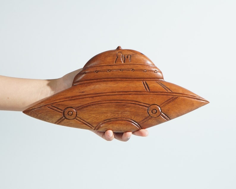 Peut inclure: Une sculpture en bois d'une soucoupe volante, avec une finition brune et des d&eacute;tails complexes. La soucoupe a un dessus arrondi et un fond plat, avec une fen&ecirc;tre circulaire de chaque c&ocirc;t&eacute;.