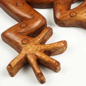 May include: A carved wooden frog sculpture featuring a smooth, polished brown finish with visible wood grain. The limbs are bent at the joints, and the feet display distinct, elongated toes. Small, circular indentations are etched into the surface.