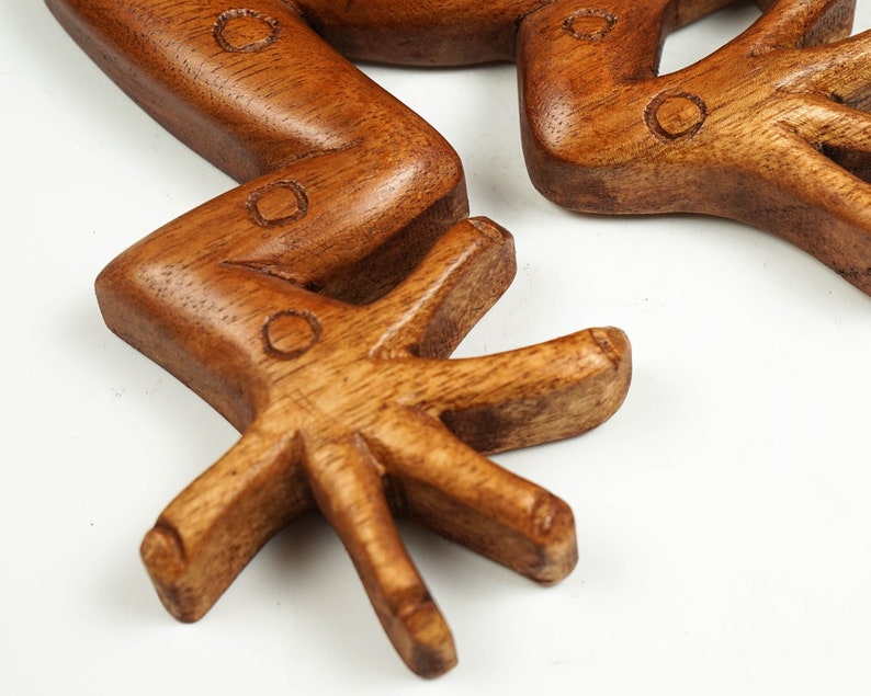 May include: A carved wooden frog sculpture featuring a smooth, polished brown finish with visible wood grain. The limbs are bent at the joints, and the feet display distinct, elongated toes. Small, circular indentations are etched into the surface.
