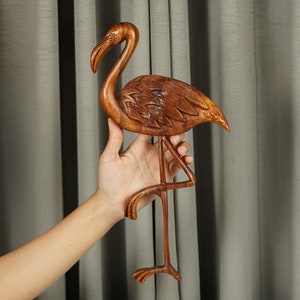 Pu&ograve; includere: Una scultura di fenicottero in legno con dettagli intricati, scolpita in legno marrone scuro. La scultura &egrave; in piedi su una sola gamba e ha la testa inclinata di lato.