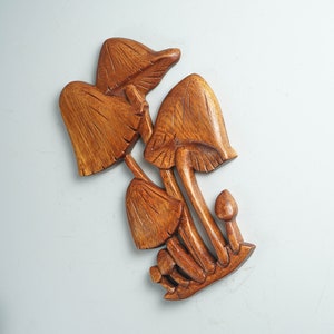 Op de afbeelding: Een handgesneden houten wanddecoratie met een groep van vijf paddenstoelen van verschillende groottes. Het beeldhouwwerk toont een rijke bruine houtnerf met gedetailleerde verticale lijnen in de hoeden en stelen van de paddenstoelen.